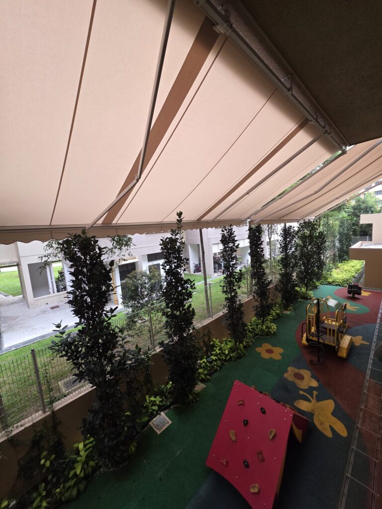 Underside of beige retractable awning fabric with articulated support arms above an outdoor playground