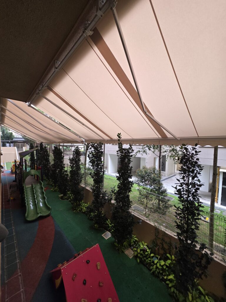 Beige retractable shade canopy covering a landscaped playground with colourful flooring and play equipment